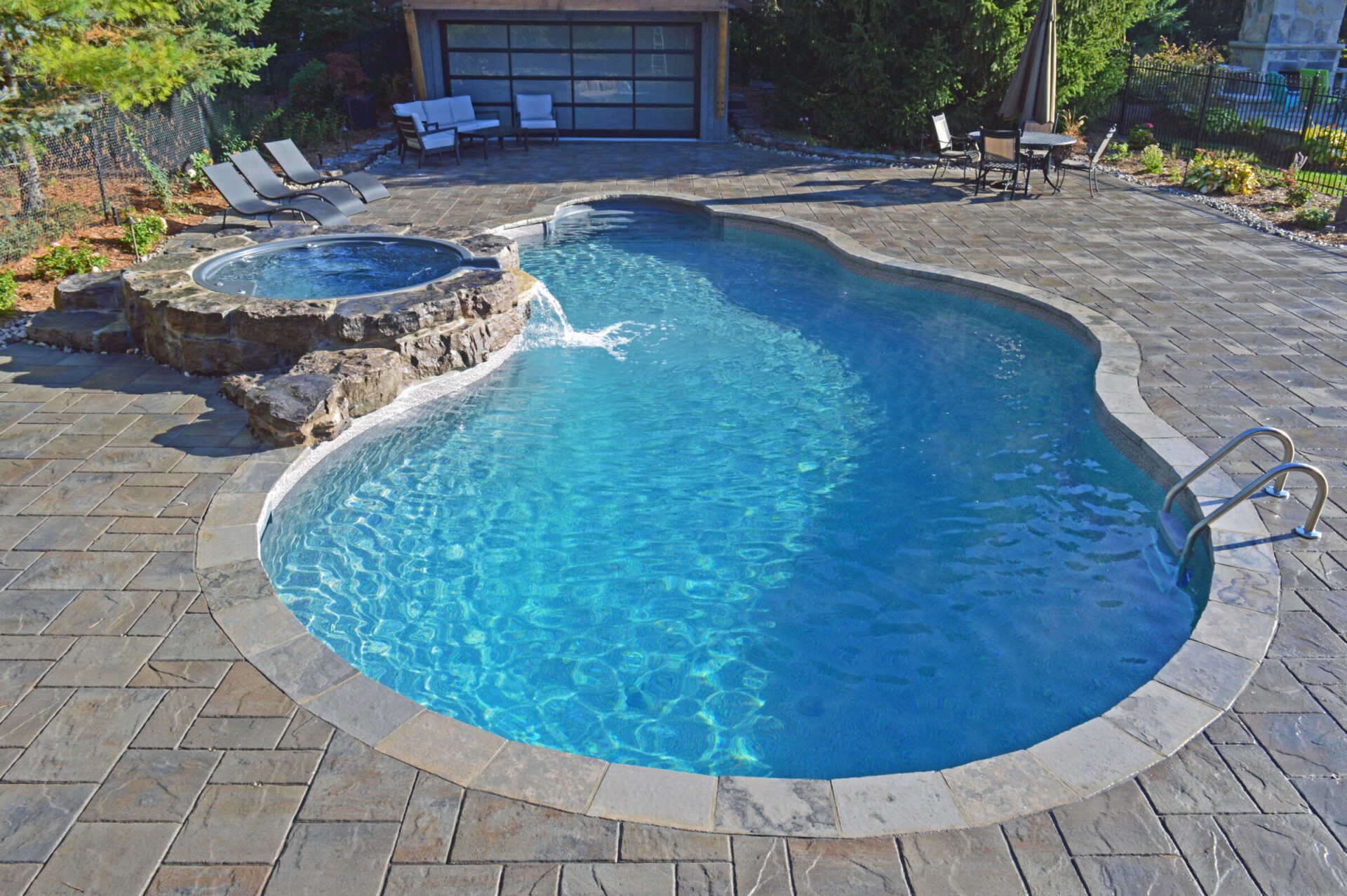 A serene backyard with a curvy pool and hot tub, surrounded by patio furniture and lush greenery, under a clear blue sky.