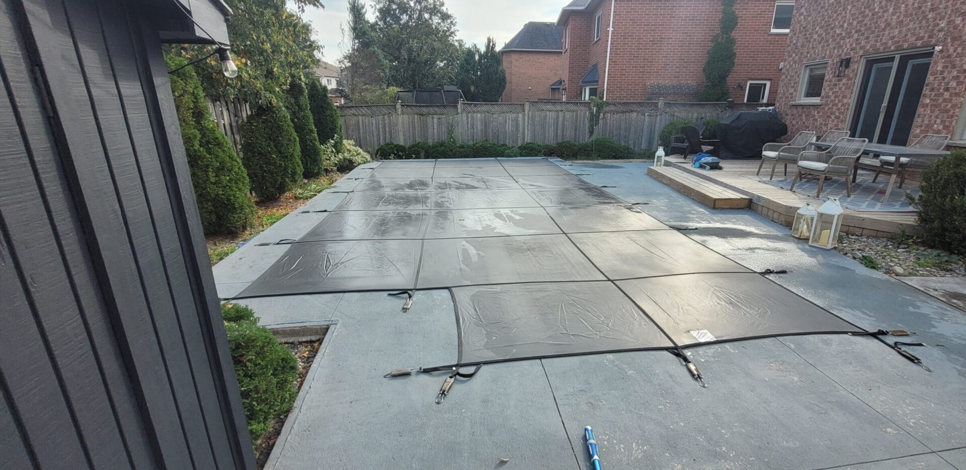 A backyard with a pool covered by a tarp, surrounded by trees, shrubs, and a wooden fence. Patio furniture is visible on the right.