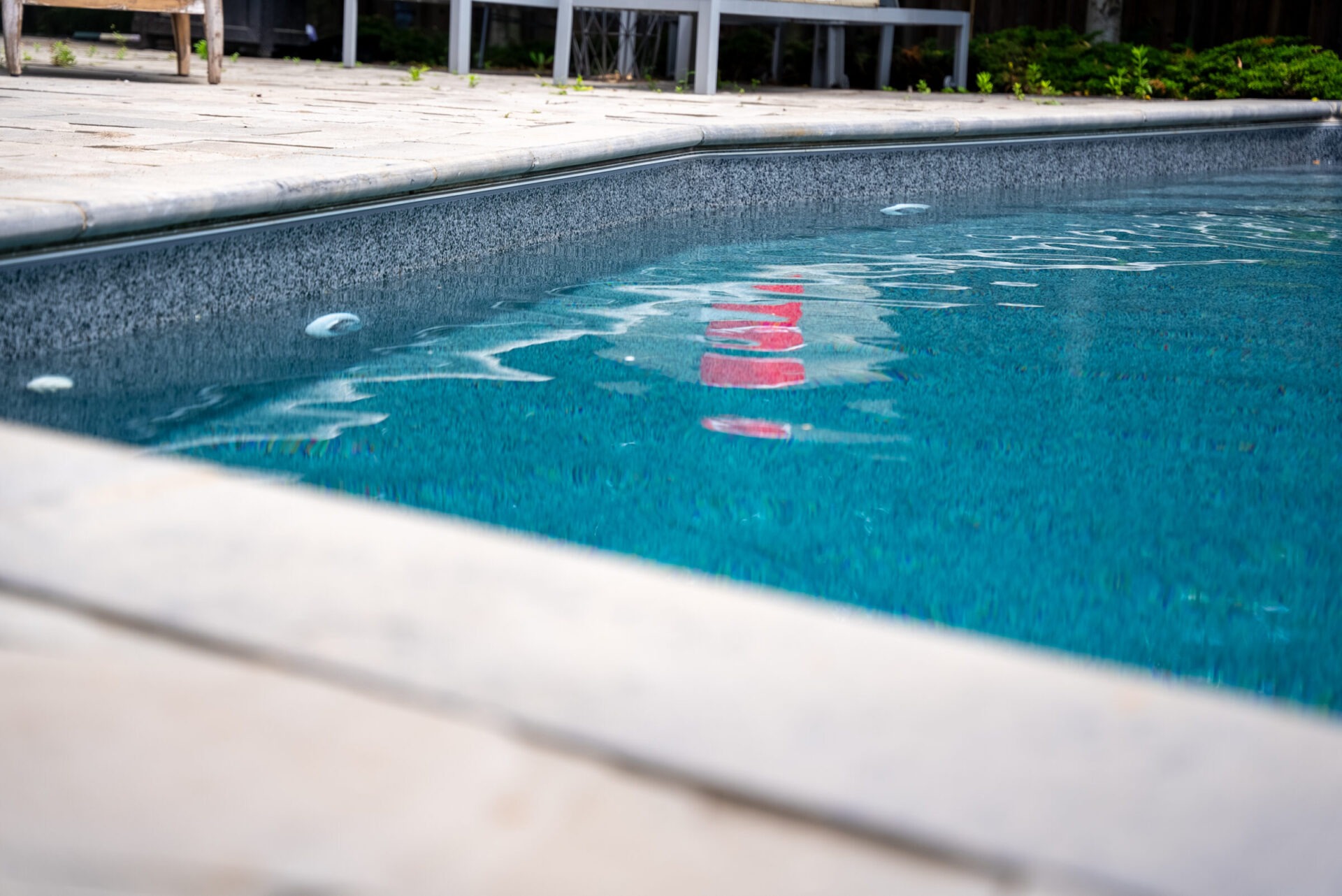 A swimming pool with clear blue water reflects red objects. Nearby is a stone patio, chairs, and a lush green garden.