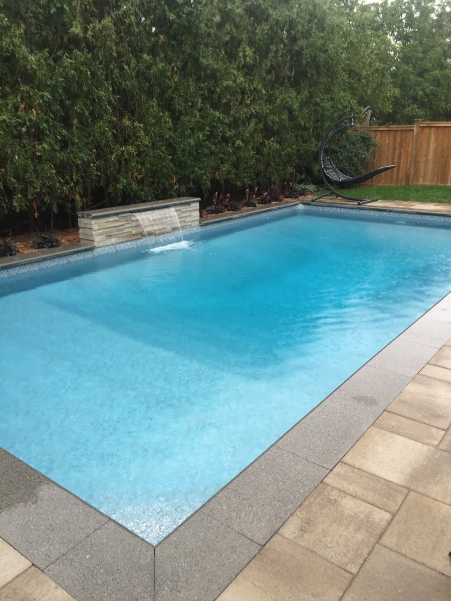 A serene backyard pool with a small waterfall feature, surrounded by lush greenery and a wooden fence, includes a hanging chair.