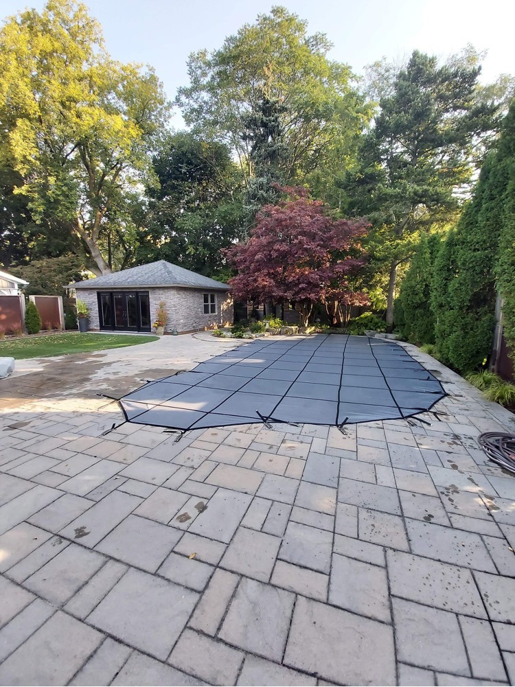 A stone patio with a covered pool, surrounded by trees and a small building with large windows in a backyard setting.