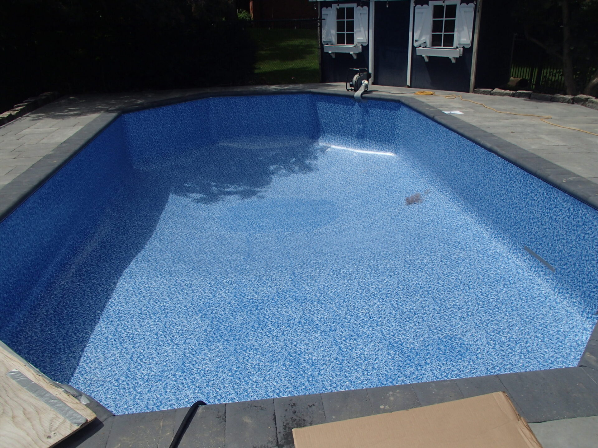 An empty residential swimming pool with a blue patterned liner, surrounded by gray tiles. A small building is visible in the background.