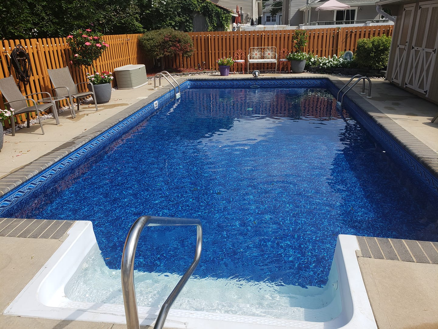 A backyard swimming pool surrounded by patio chairs, potted plants, and a wooden fence on a sunny day. No people are present.