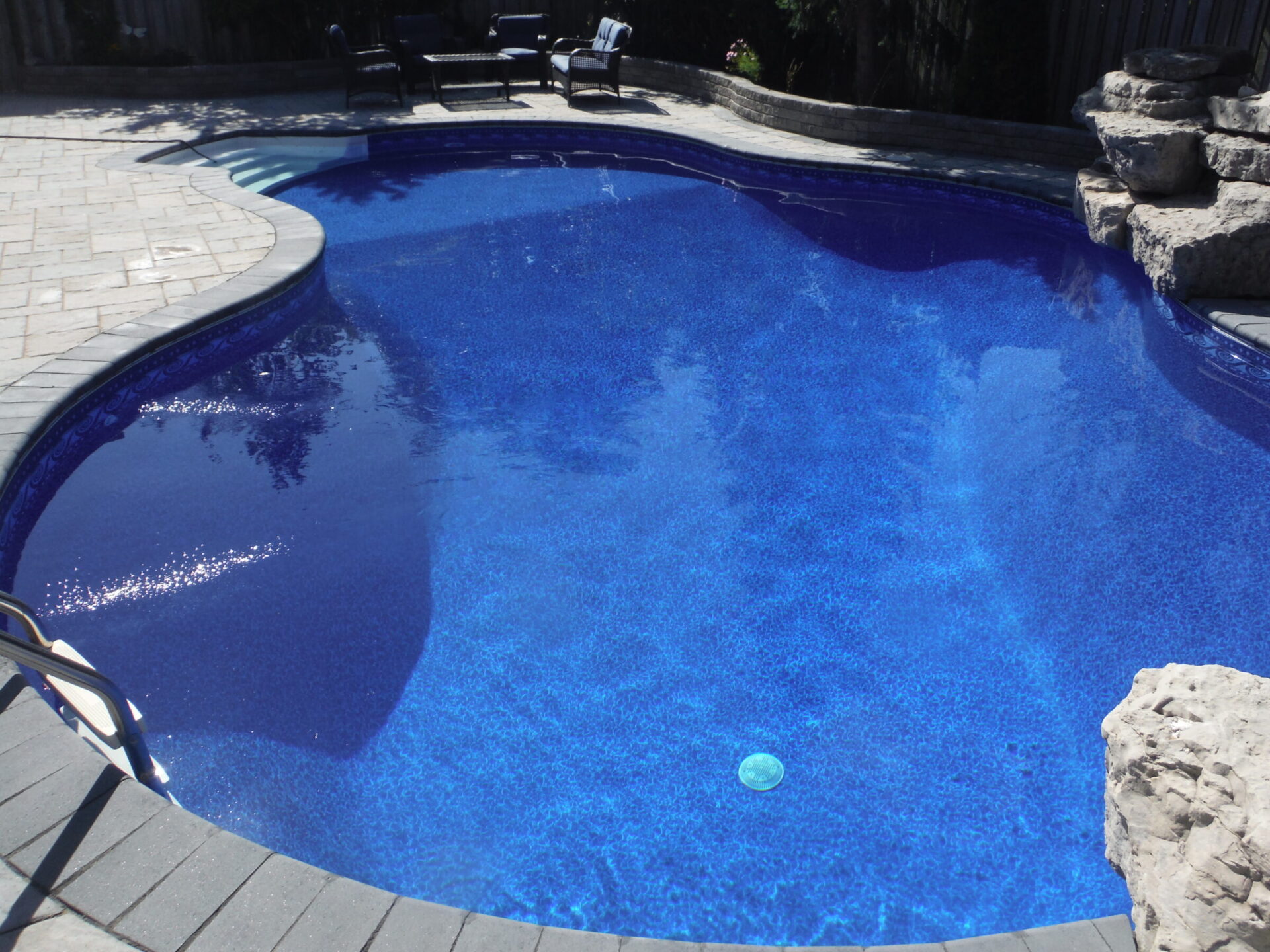 A clear blue swimming pool surrounded by stone tiles and rocks, with outdoor chairs in the background, under bright sunlight.