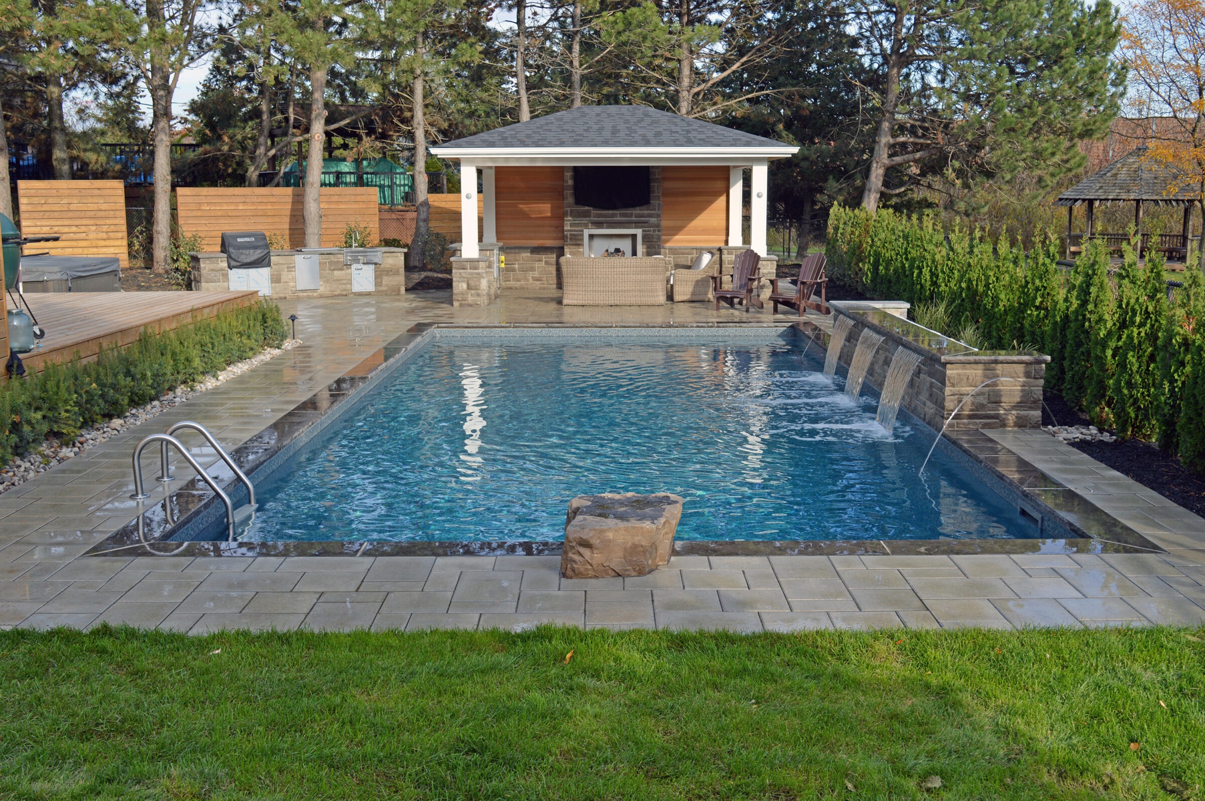 A serene backyard scene with a rectangular pool, waterfall feature, and patio. Surrounded by trees and shrubs, beneath a clear sky.