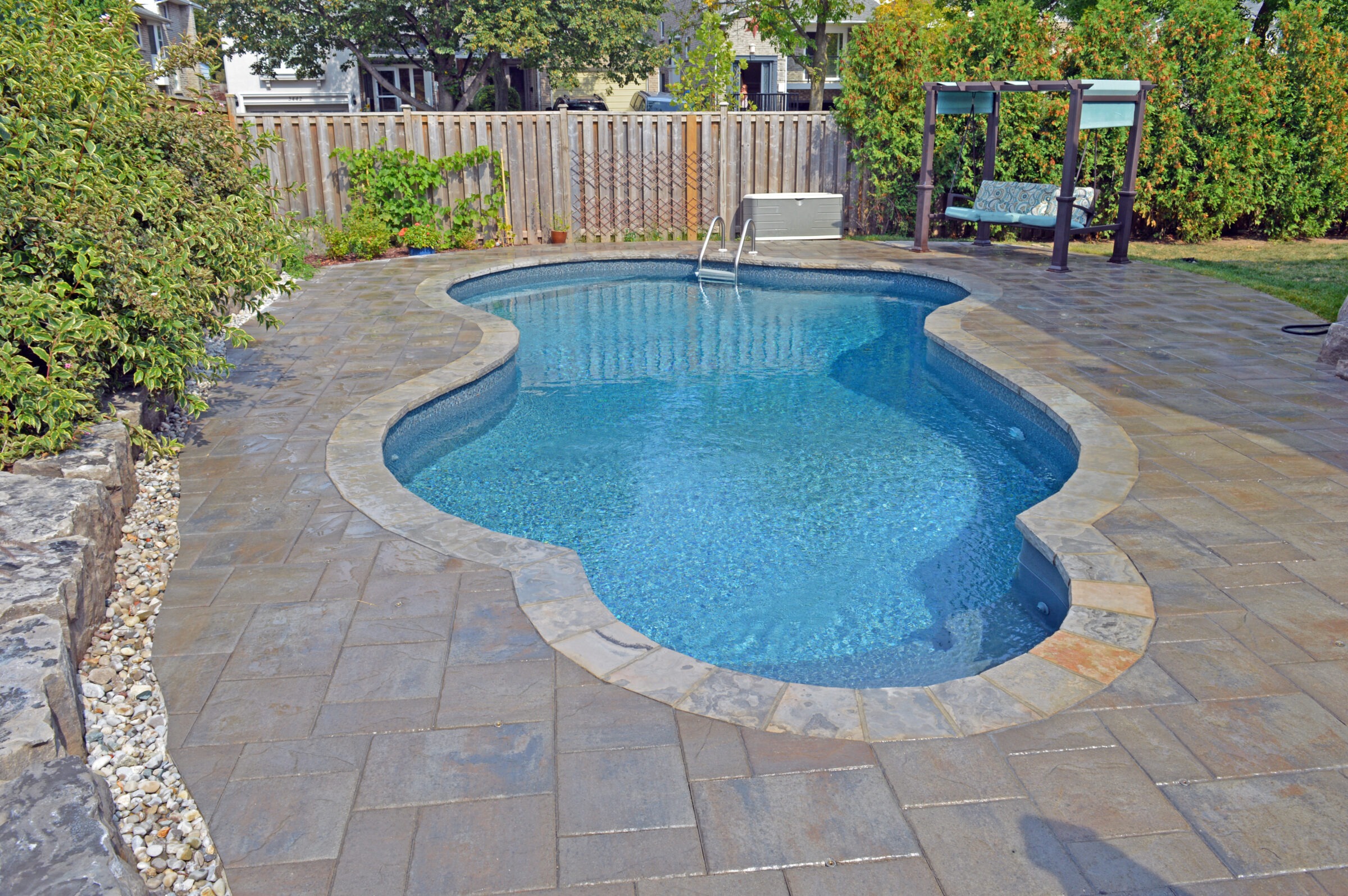 A small backyard pool with stone patio, wooden fence, and surrounding greenery. A swing set is visible in the corner. Quiet and serene.