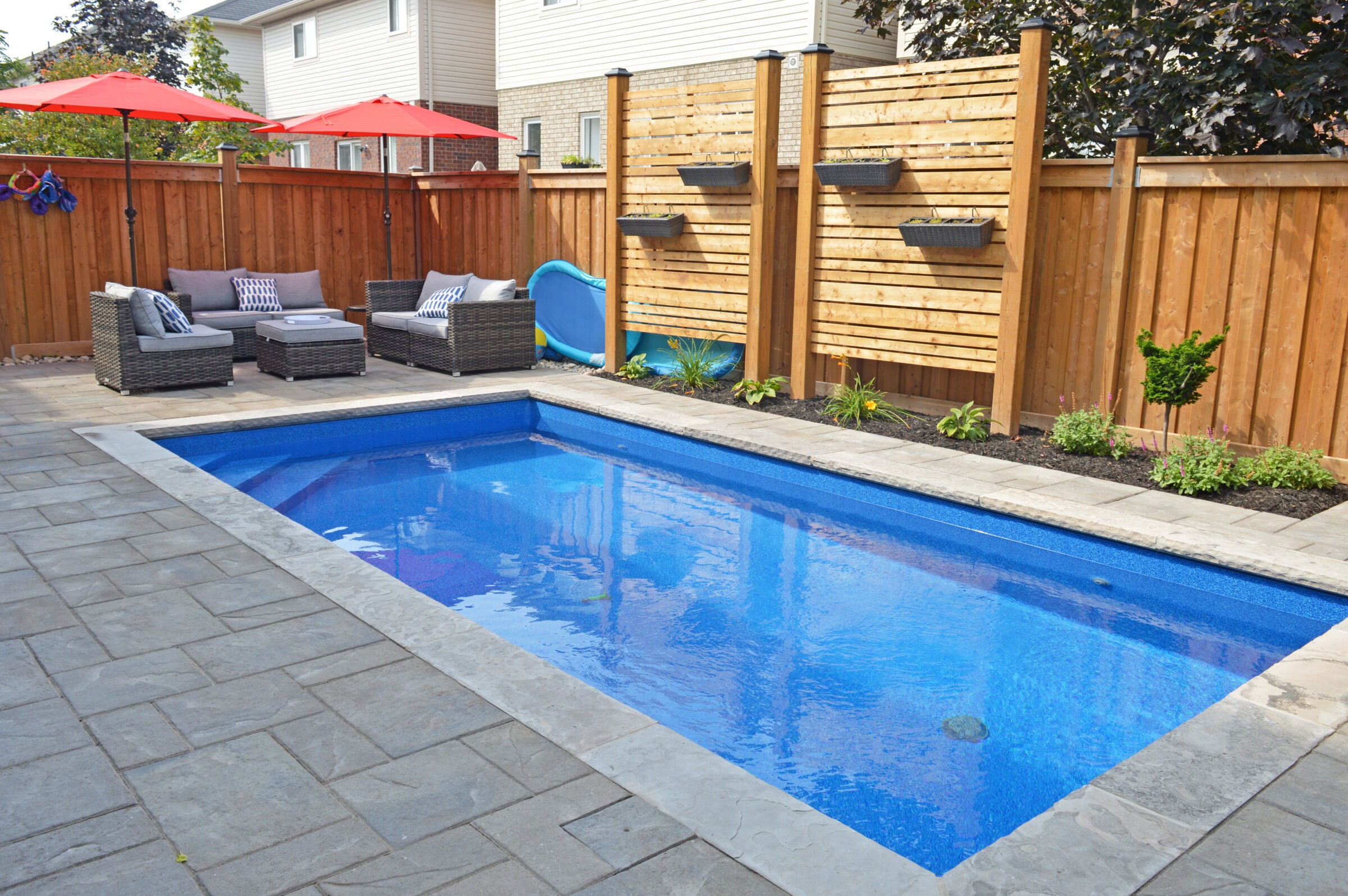 A residential backyard features a rectangular pool, patio with seating, and a wooden fence. Red umbrellas provide shade over the lounge area.