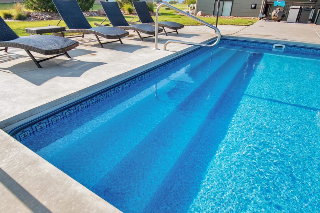 A sparkling outdoor pool with several lounge chairs. Bright sunlight enhances the serene setting. A small yard and building are visible in the background.