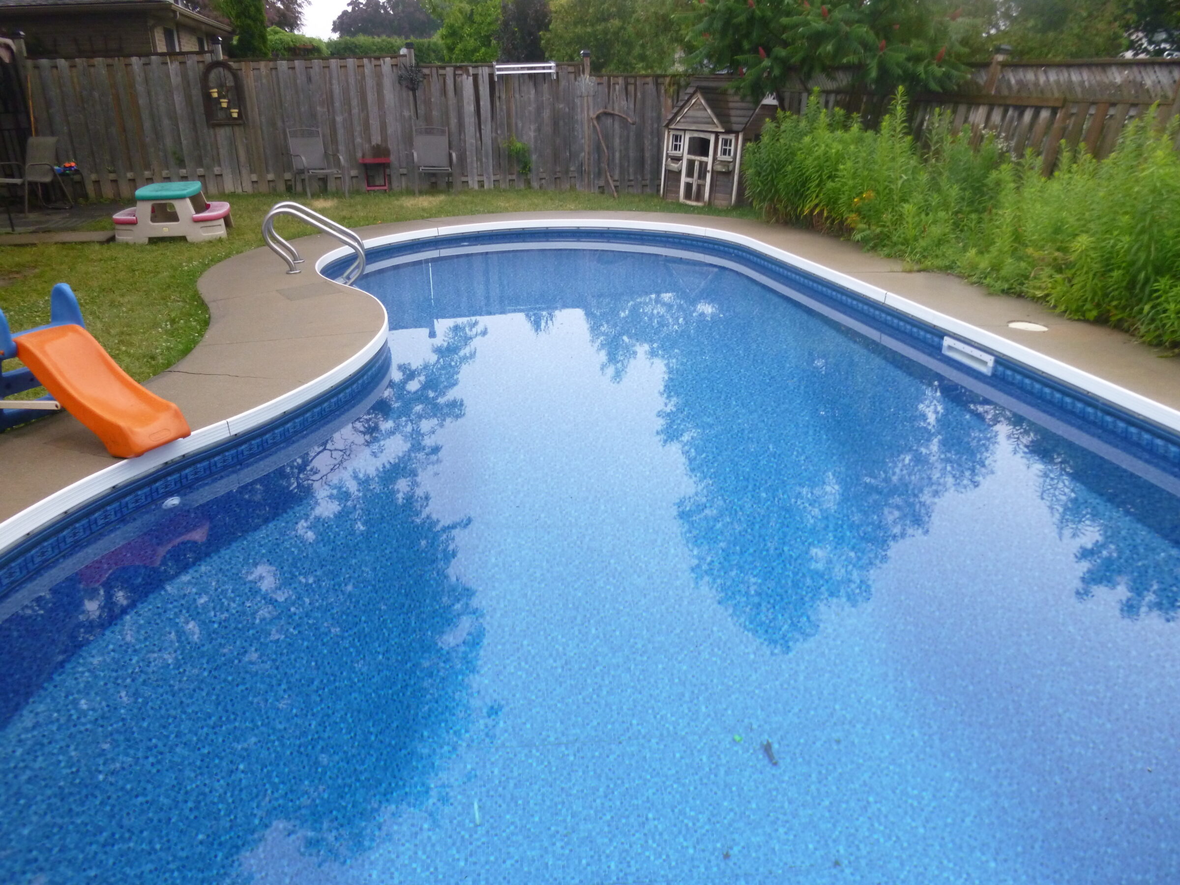 A backyard with a curved swimming pool, surrounded by grass and a fence. A small slide and kid's playhouse are visible.