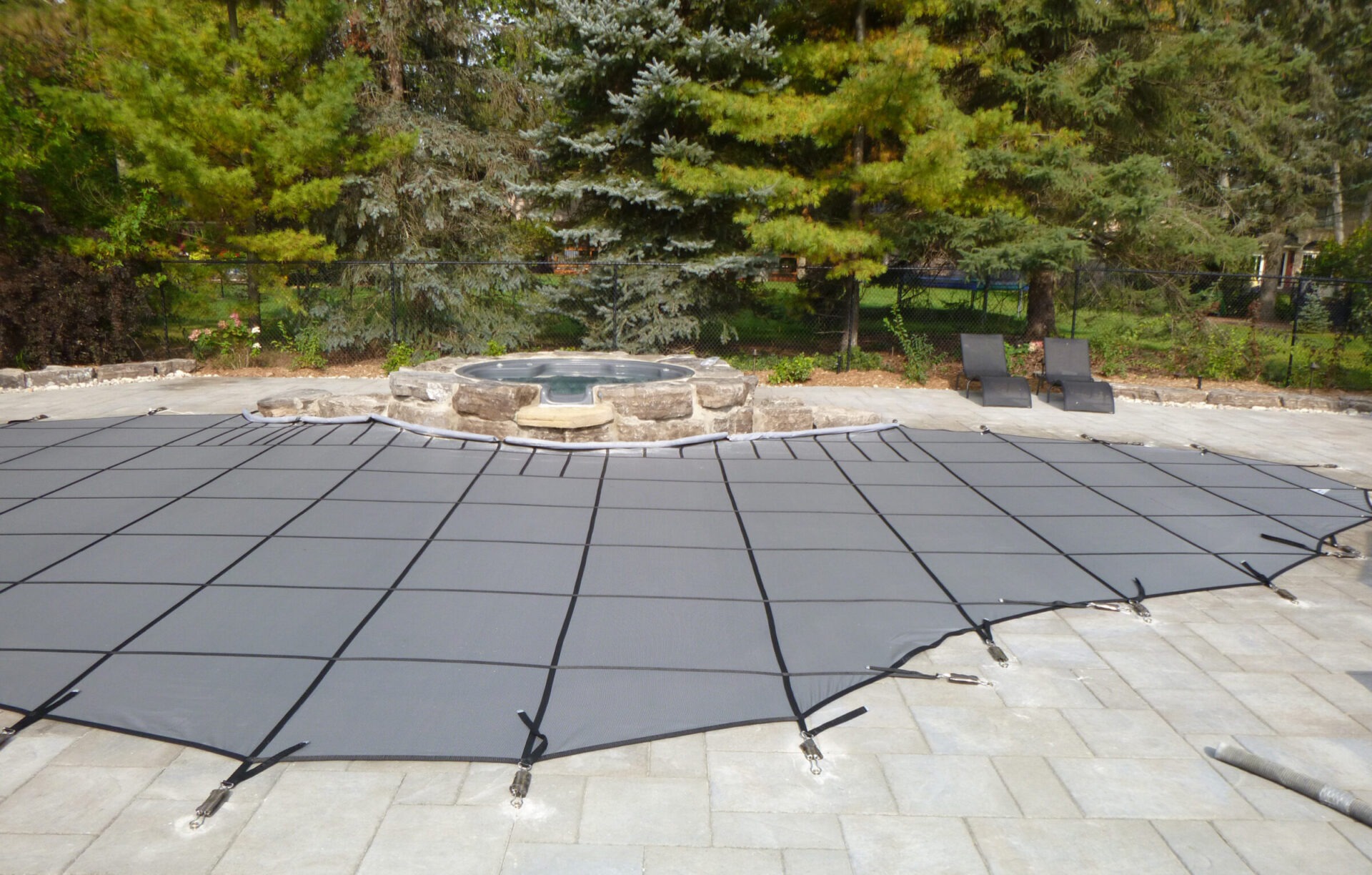 A covered pool with a safety cover, surrounded by trees and a stone patio. Two lounge chairs sit near a small, round spa.