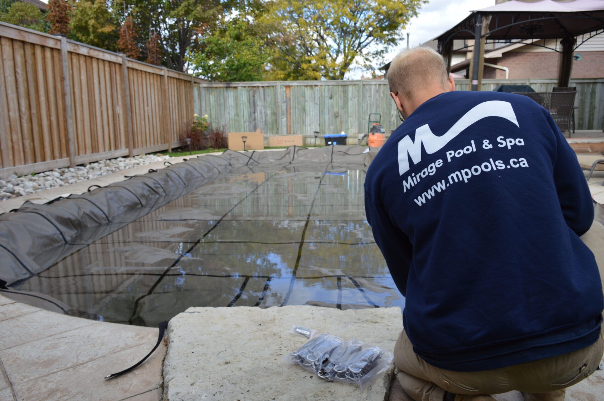 A person with "Mirage Pool & Spa" shirt works on winterizing a backyard pool, surrounded by a wooden fence and trees.