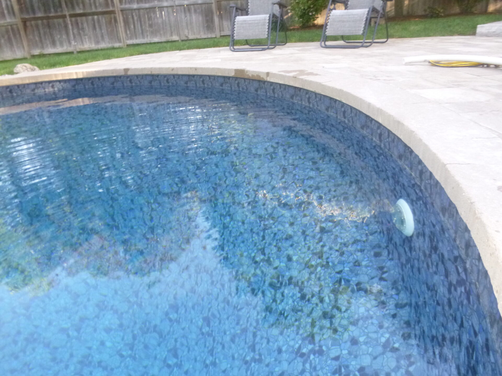 A serene outdoor pool with clear blue water, surrounded by a stone patio and patio chairs, next to a wooden fence.