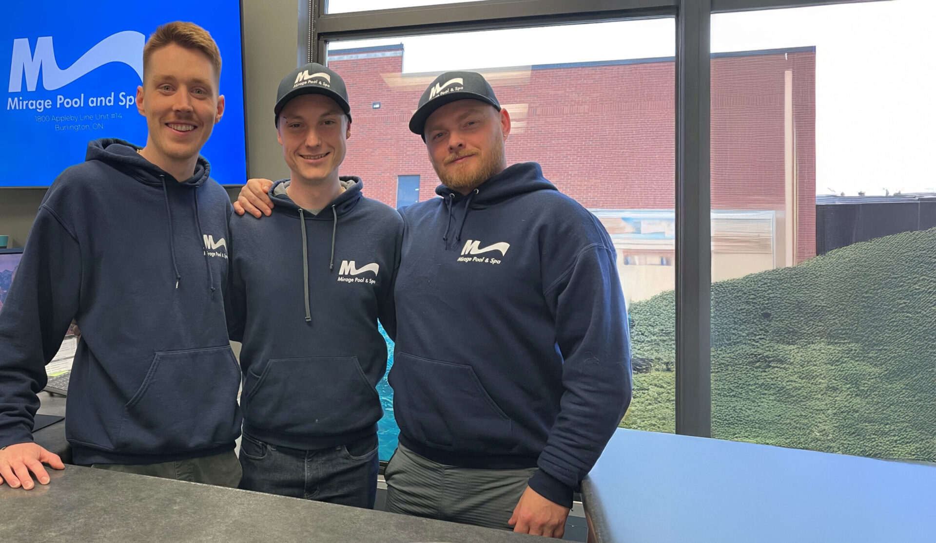 Three people wearing Mirage Pool and Spa hoodies stand together indoors, smiling. A large window reveals an outdoor view with a brick building.