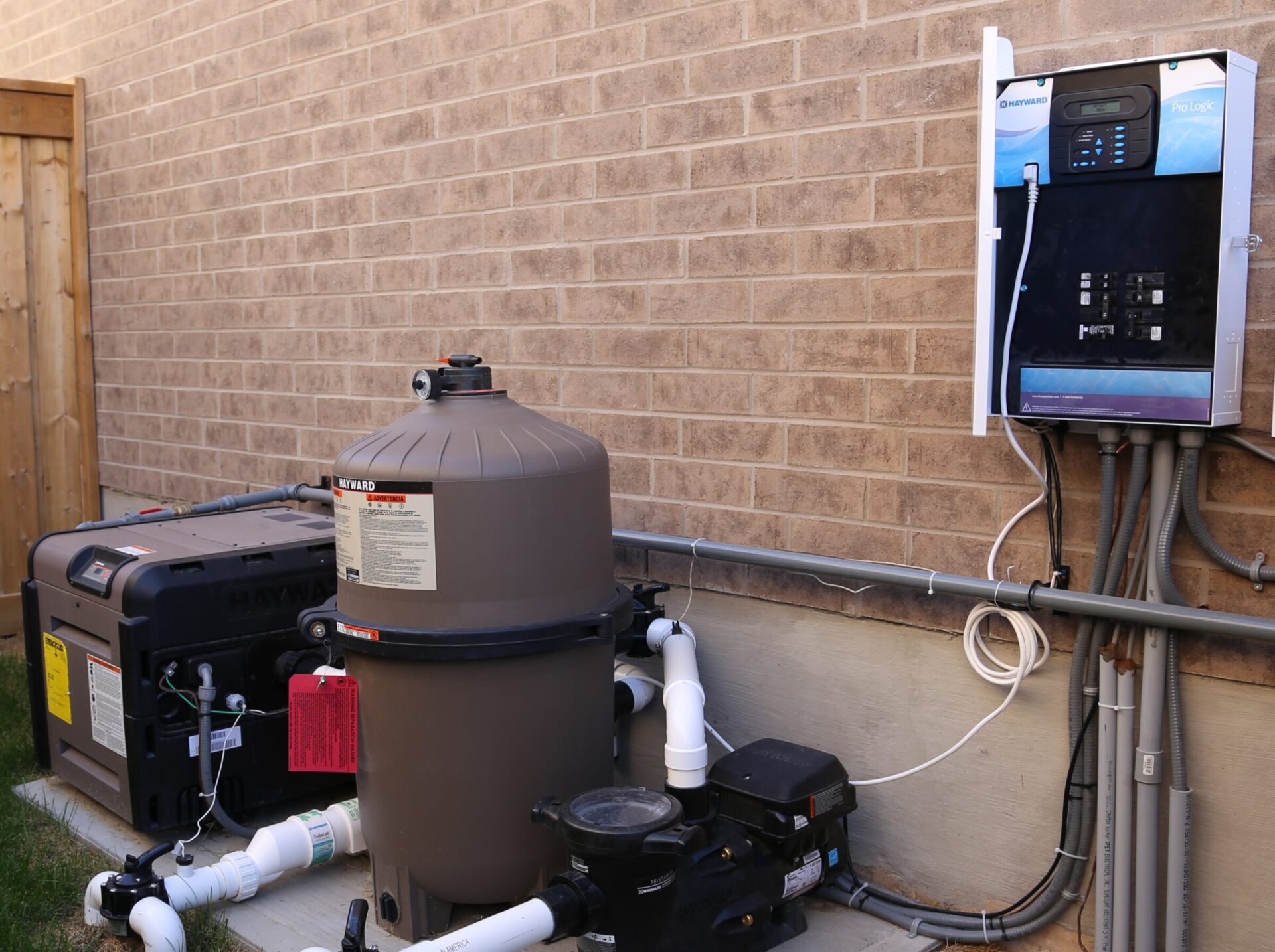 Pool equipment setup outdoors includes a sand filter, heater, and control panel mounted on a brick wall, connected with pipes and wires.